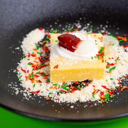 Close-up of a slice of cake with cream and cherry on a black plate, decorated with colorful sprinkles.