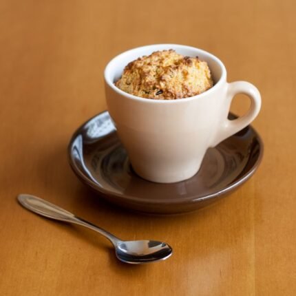Warm mug cake in a white cup on a brown saucer with a spoon on a wooden table.